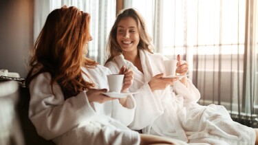 Zwei Frauen in Bademänteln trinken Kaffee oder Tee und lächeln sich in entspannter Atmosphäre an