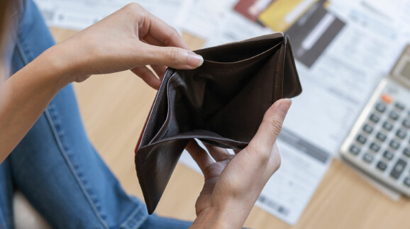 Eine Person hält eine leere Geldbörse in den Händen. Im Hintergrund liegen Rechnungen, Kreditkarten und ein Taschenrechner auf dem Tisch.