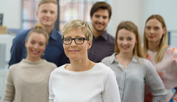 Eine Frau mit Brille steht im Vordergrund, im Hintergrund stehen f&uuml;nf weitere Personen, die leicht unscharf sind. Alle l&auml;cheln in die Kamera.