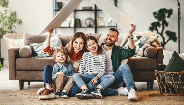 Vierköpfige Familie sitzt lachend auf dem Wohnzimmerboden und hält ein symbolisches Dach aus Holz über ihren Köpfen.