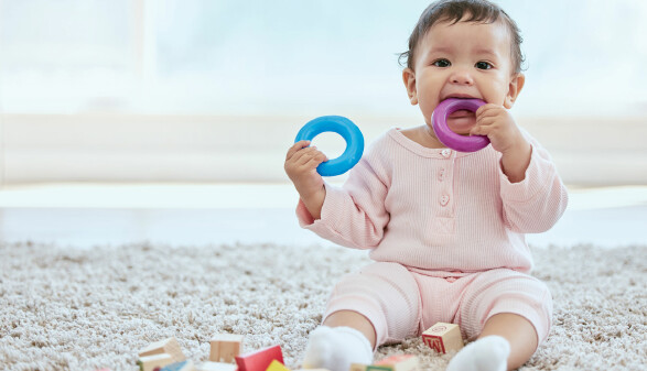 Baby sitzt am Boden, hat einen blauen Silikonring in der Hand und einen violetten im Mund.