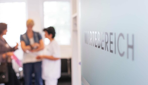 Zwei Patientinnen unterhalten sich mit einer Ordinationsgehilfin. Rechts ist auf einer wei&szlig;en Wand Wartebereich zu lesen.