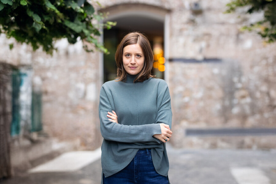 Claudia Endrich steht mit verschränkten Armen vor einem Steinbau. Sie trägt einen graublauen Pullover und blickt ruhig in die Kamera.