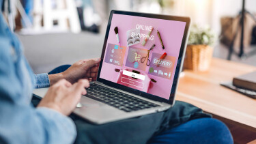 Person sitzt auf dem Sofa und nutzt einen Laptop, auf dem eine Online-Shopping-Seite mit Kosmetikartikeln und Anmeldefeldern angezeigt wird.