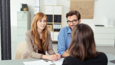 Junges Paar sitzt in einem Büro an einem Tisch und spricht freundlich mit einer Beraterin, auf dem Tisch liegen Dokumente.