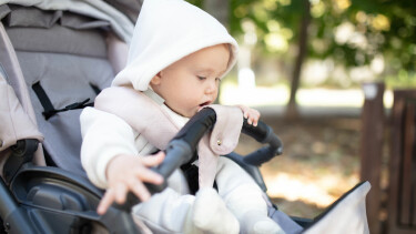 Baby in einem Kinderwagen mit Kapuzenpullover, das neugierig die Sicherheitsstange betrachtet und mit der Hand danach greift.