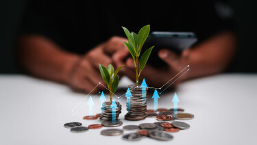 Münzstapel mit wachsenden grünen Pflänzchen, davor verstreute Münzen; im Hintergrund unscharfe Person mit Smartphone. Grafische Pfeile und Linien symbolisieren finanzielles Wachstum.