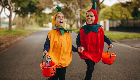 Zwei fröhliche Kinder in bunten Halloweenkostümen laufen lachend Hand in Hand eine Straße entlang und tragen orangene Eimer zum Süßigkeitensammeln.