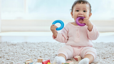 Baby sitzt am Boden, hat einen blauen Silikonring in der Hand und einen violetten im Mund.