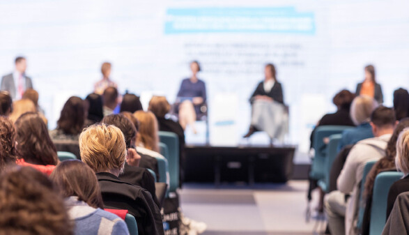 Teilnehmerinnen und Teilnehmer verfolgen eine Podiumsdiskussion auf einer Konferenz mit mehreren Personen auf der Bühne.