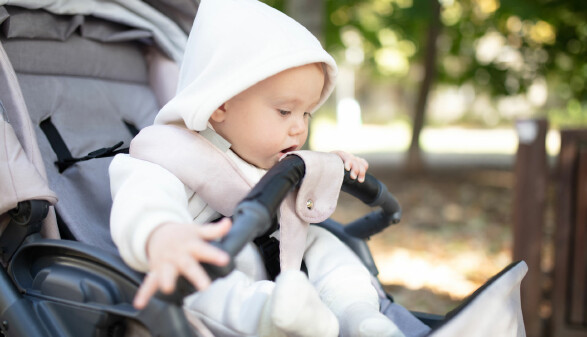 Baby in einem Kinderwagen mit Kapuzenpullover, das neugierig die Sicherheitsstange betrachtet und mit der Hand danach greift.