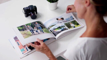 Eine Frau betrachtet ein geöffnetes Fotobuch mit Familienfotos auf einem weißen Tisch, daneben liegen eine Kamera und eine kleine Pflanze.
