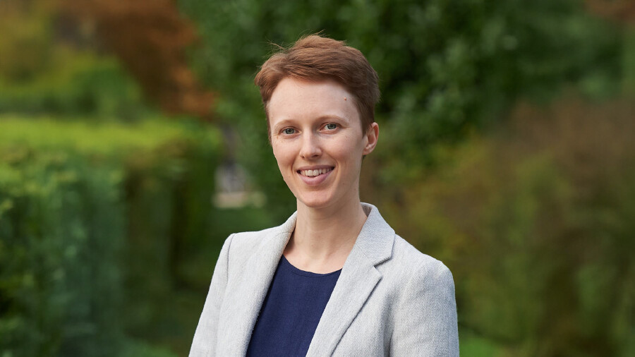 Anna Silber ist im Porträt zu sehen. Sie steht im Freien vor einer grünen, leicht verschwommenen Naturkulisse. Sie trägt einen hellgrauen Blazer und ein dunkelblaues Oberteil. Ihr kurzer, rötlich-brauner Haarschnitt ist ordentlich frisiert. Sie blickt freundlich in die Kamera.