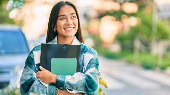 Im Vordergrund steht eine junge Person im Freien, die lächelnd zur Seite blickt. Sie hält mehrere Unterlagen und Hefte im Arm und trägt eine grün-karierte Jacke. Im Hintergrund ist eine unscharfe Straßen- bzw. Gehwegsituation mit Grünpflanzen zu sehen. Die Umgebung wirkt freundlich und sonnig. © krakenimages.com, stock.adobe.com Junge Person im Freien, lächelnd und mit Ordnern und Heften im Arm.