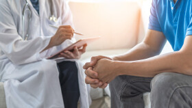 &Auml;rztin mit umgeh&auml;ngtem Stethoskop sitzt, macht sich Notizen auf einem IPad. Daneben sitzt ein Patient mit blauem Poloshirt