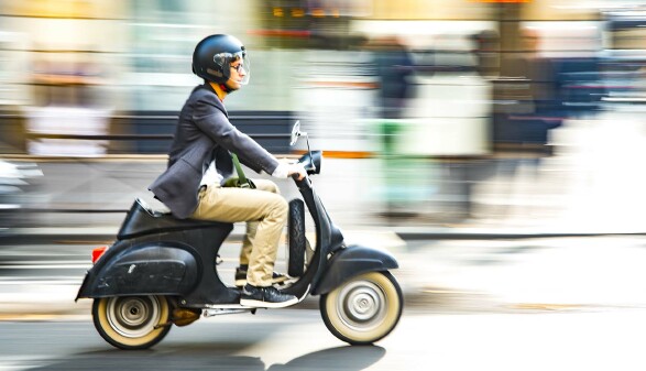 Ein junger Mann mit schwarzem Helm f&auml;hrt mit dem Moped durch die Stadt.