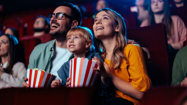 Eine Familie - Vater, Mutter, Tochter - sitzt mit 2 gro&szlig;en T&uuml;ten Popcorn im Kinosaal und hat sichtlich Freude am Film.