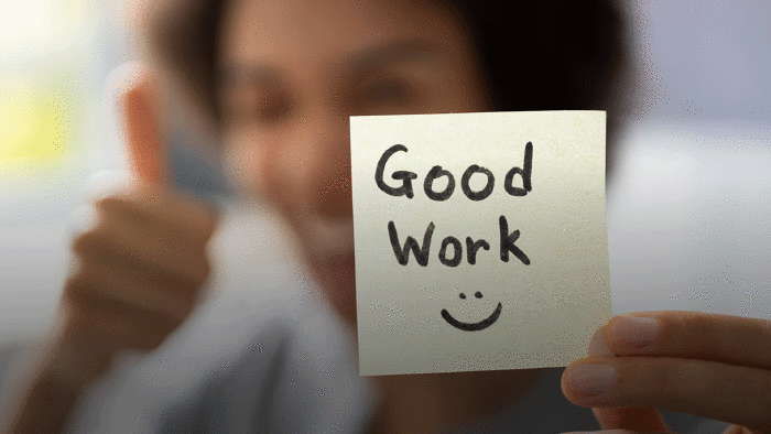 Person h&auml;lt einen Zettel mit der Aufschrift &bdquo;Good Work&ldquo; und zeigt Daumen hoch.