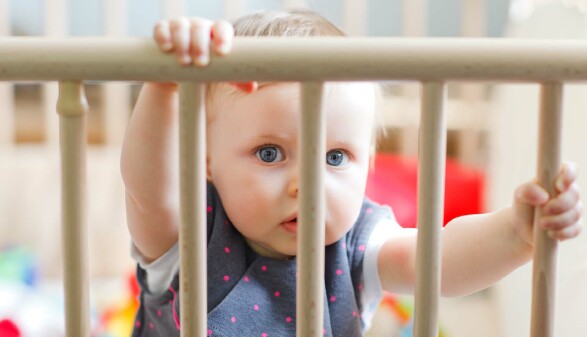 Baby hält sich mit beiden Händen an einem Treppenschutz fest und schaut neugierig hindurch.