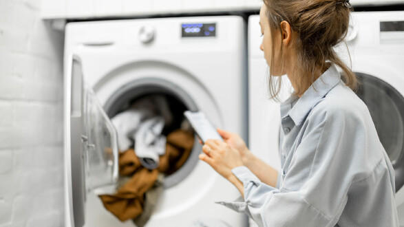 Frau bel&auml;dt eine Waschmaschine und pr&uuml;ft auf ihrem Smartphone Waschprogramm und Energieverbrauch im modernen Waschraum.