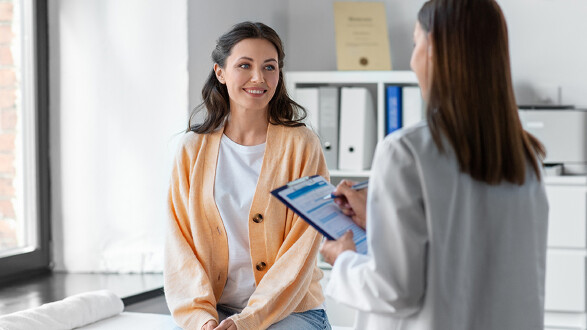 Eine Patientin sitzt lächelnd in einem Untersuchungsraum, während eine Ärztin ihr gegenübersteht und Notizen auf einem Klemmbrett macht. © Syda Productions, stock.adobe.com Eine Patientin sitzt lächelnd in einem Untersuchungsraum, während eine Ärztin ihr gegenübersteht und Notizen auf einem Klemmbrett macht.
