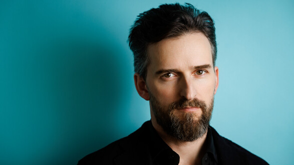 Porträt eines Mannes mit dunklem Haar und vollem Bart vor einem blaugrünen Hintergrund.