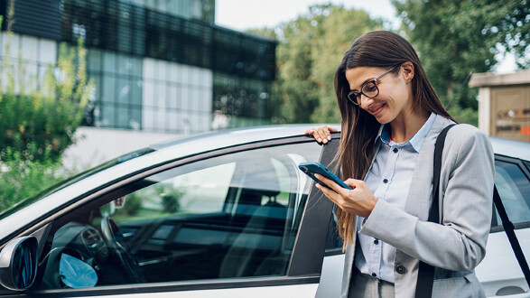 Eine Frau in Businesskleidung steht neben einem Auto und schaut lächelnd auf ihr Smartphone. © Zamrznuti tonovi, stock.adobe.com Eine Frau in Businesskleidung steht neben einem Auto und schaut lächelnd auf ihr Smartphone.