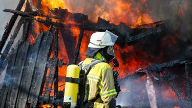 Feuerwehrkraft in Schutzkleidung mit Atemger&auml;t steht vor einem stark brennenden Geb&auml;ude mit Flammen und verkohlten Holzbalken.