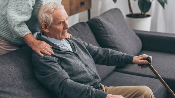 Ein alter Mann mit Gehstock in der Hand sitzt auf einem Sofa und schaut ins Leere. Eine alte Frau greift ihm von hinten auf die Schultern.