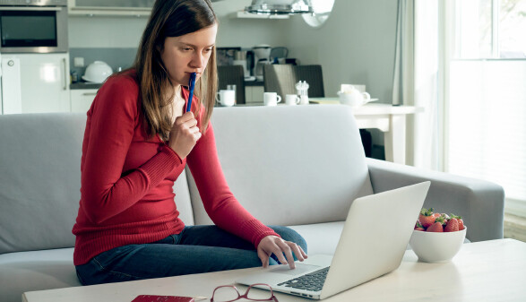 Frau sitzt auf Sofa, arbeitet konzentriert am Laptop, hält Stift in der Hand, helle moderne Wohnung.