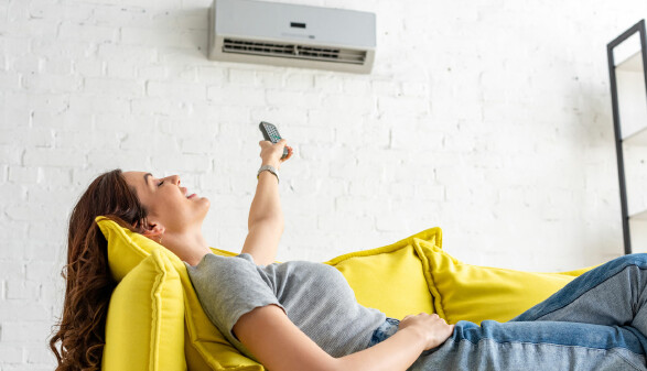 Eine junge Frau liegt entspannt auf einem leuchtend gelben Sofa und richtet eine Fernbedienung auf eine Klimaanlage an der Wand über ihr.