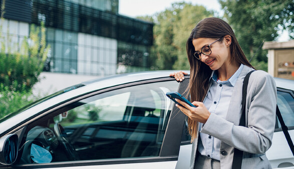Eine Frau in Businesskleidung steht neben einem Auto und schaut l&auml;chelnd auf ihr Smartphone.