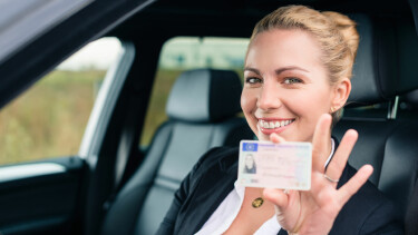 Eine fr&ouml;hliche junge Frau sitzt im Auto und zeigt ihren F&uuml;hrerschein.