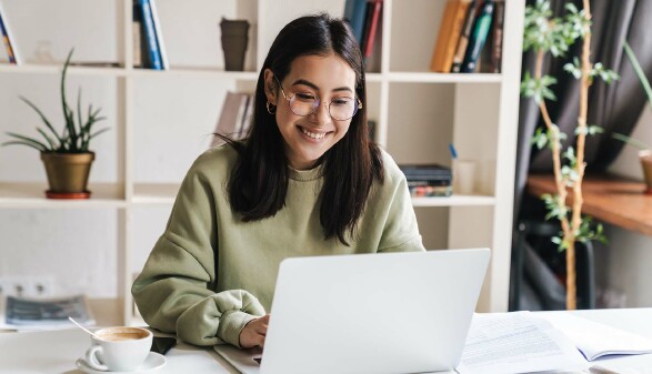 Eine Frau vor einem Laptop. Neben ihr liegen Papiere und Unterlagen. 