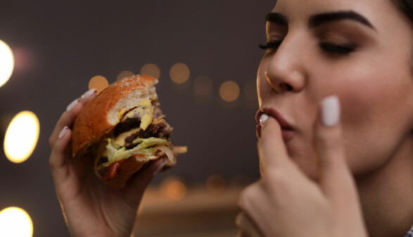 Junge Frau mit einem Burger in der Hand schleckt sich den Zeigefinger ab.