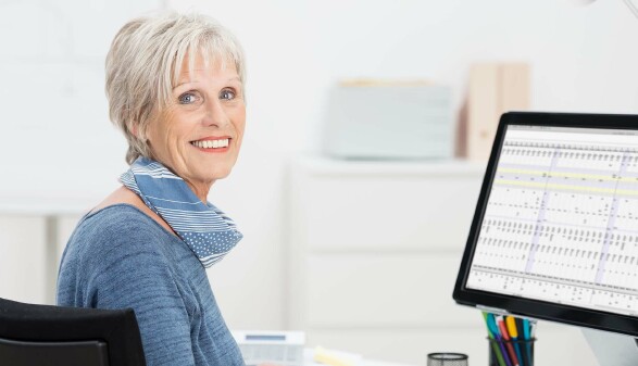 Ältere Frau mit kurzem, grauem Haar sitzt lächelnd an einem Schreibtisch und blickt zur Kamera; neben ihr steht ein Computerbildschirm mit einer geöffneten Tabellenansicht.