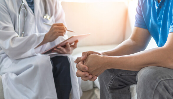 &Auml;rztin mit umgeh&auml;ngtem Stethoskop sitzt, macht sich Notizen auf einem IPad. Daneben sitzt ein Patient mit blauem Poloshirt