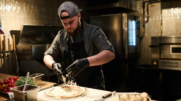 Ein Pizzabäcker belegt in einer professionellen Küche eine Pizza mit geriebenem Käse.