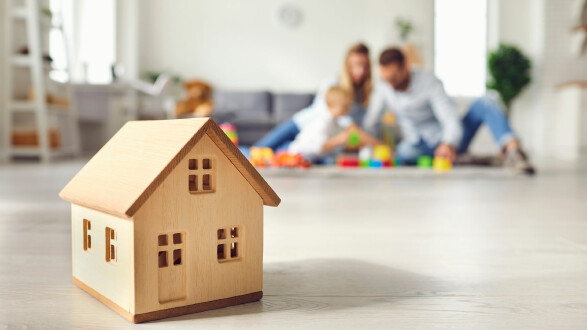 Im Vordergrund steht ein kleines, aus hellem Holz gefertigtes Modellhaus auf einem hellen Boden. Im Hintergrund ist unscharf eine Familie mit zwei Erwachsenen und einem Kleinkind zu sehen, die gemeinsam auf dem Boden im Wohnzimmer mit buntem Spielzeug spielen. Der Raum wirkt hell und freundlich, mit Pflanzen und modernen Möbeln. © Studio Romantic, shutterstock.com Holzspielzeughaus im Vordergrund, während im Hintergrund eine Familie mit einem Kind unscharf auf dem Boden spielt.