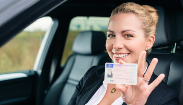 Eine fr&ouml;hliche junge Frau sitzt im Auto und zeigt ihren F&uuml;hrerschein.