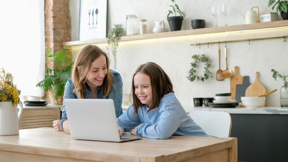 Das Bild zeigt eine helle, gemütliche Küche mit modernen und natürlichen Elementen. Links im Vordergrund sitzt eine Frau mit schulterlangem, hellbraunem Haar in einem blauen Jeanshemd. Neben ihr sitzt ein Mädchen mit braunem, schulterlangem Haar in einem blau-weiß gestreiften Langarmshirt. Beide schauen lächelnd auf einen Laptop auf dem Tisch. Die Küche im Hintergrund ist mit Pflanzen, Küchenutensilien und Dekoobjekten wie Schneidebrettern und Gläsern gestaltet. Die Szene wirkt freundlich, häuslich und zeigt ein gemeinsames, positives Erlebnis – möglicherweise beim Lernen oder Spielen am Laptop. © lelechka, stock.adobe.com Eine Frau und ein Mädchen sitzen gemeinsam an einem Holztisch in einer hellen, modern eingerichteten Küche. Sie schauen lächelnd auf einen aufgeklappten Laptop. Die Atmosphäre ist freundlich und entspannt.