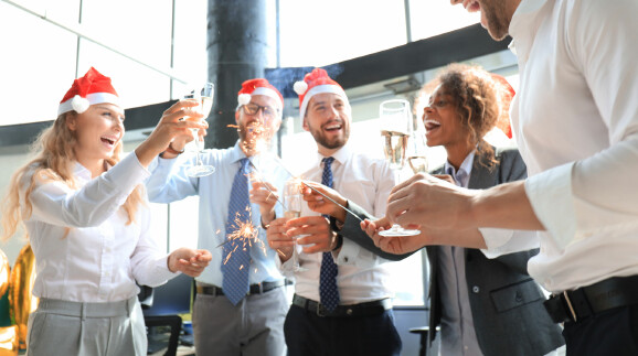 Gruppe von Kolleginnen und Kollegen feiert fröhlich im Büro mit Sektgläsern, Wunderkerzen und Weihnachtsmützen.
