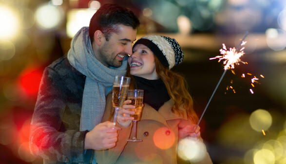 L&auml;chelndes Paar st&ouml;&szlig;t an Silvester mit Sekt an, Frau h&auml;lt Wunderkerze in winterlicher Abendstimmung.