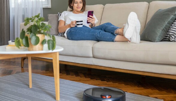Ein Saugroboter vor einer Couch, auf der eine junge Frau liegt, eine Kaffeetasse in der Hand h&auml;lt und auf ihr Handy schaut