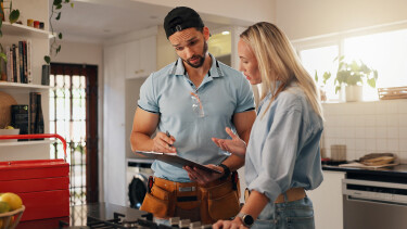 Ein Installateur und eine Frau stehen in einer hellen Küche und besprechen etwas auf einem Klemmbrett.
