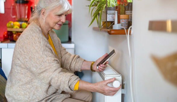 &Auml;ltere Frau h&auml;lt ein Handy in der Hand, w&auml;hrend sie das Thermostat am Heizk&ouml;rper zu Hause einstellt