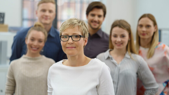 Das Bild zeigt eine Frau mittleren Alters mit kurzen, blonden Haaren und schwarzer Brille im Vordergrund. Sie trägt ein schlichtes, weißes Oberteil und lächelt dezent in die Kamera. Im Hintergrund stehen fünf weitere Personen – drei Frauen und zwei Männer – die ebenfalls lächeln, jedoch leicht unscharf abgebildet sind. Die Gruppe befindet sich in einem modernen, hellen Büro mit Tageslicht. Die Stimmung wirkt freundlich und professionell, was auf ein Team- oder Unternehmensporträt hinweist. © contrastwerkstatt, stock.adobe.com Eine Frau mit Brille steht im Vordergrund, im Hintergrund stehen fünf weitere Personen, die leicht unscharf sind. Alle lächeln in die Kamera.