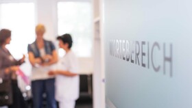 Zwei Patientinnen unterhalten sich mit einer Ordinationsgehilfin. Rechts ist auf einer wei&szlig;en Wand Wartebereich zu lesen.