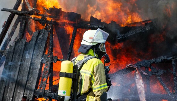 Feuerwehrkraft in Schutzkleidung mit Atemger&auml;t steht vor einem stark brennenden Geb&auml;ude mit Flammen und verkohlten Holzbalken.