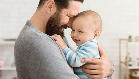 Vater h&auml;lt l&auml;chelnd sein Baby im Arm; beide ber&uuml;hren lachend die Stirn aneinander.